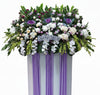 A large floral condolence arrangement in a white and purple draped basket, featuring white orchids, purple and white roses, carnations, and green foliage, with a 'Condolences' sign in the center, shown against a plain white background.
