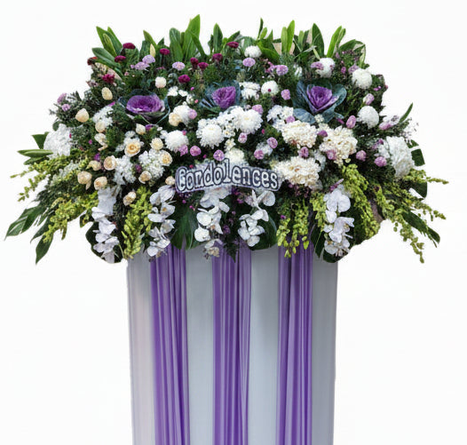 A large floral condolence arrangement in a white and purple draped basket, featuring white orchids, purple and white roses, carnations, and green foliage, with a 'Condolences' sign in the center, shown against a plain white background.