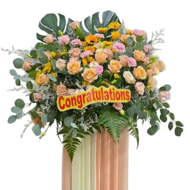 A tall floral arrangement on a striped pedestal with pastel peach, cream, and pale green panels. The bouquet includes pink and yellow roses, green foliage, eucalyptus leaves, and a yellow sign reading 'Congratulations' against a plain indoor wall.