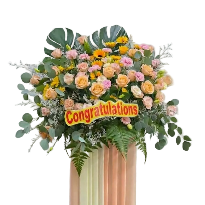 A tall floral arrangement on a striped pedestal with pastel peach, cream, and pale green panels. The bouquet includes pink and yellow roses, green foliage, eucalyptus leaves, and a yellow sign reading 'Congratulations' against a plain indoor wall.