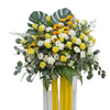 A vibrant floral arrangement featuring yellow and white roses, eucalyptus, monstera leaves, and ferns, displayed on a white stand with two vertical yellow ribbons.