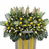 A large floral arrangement for condolence, featuring yellow and white flowers, green foliage, and tall yellow drapes on a white and yellow stand, with a sign reading 'Deepest Condolence'.